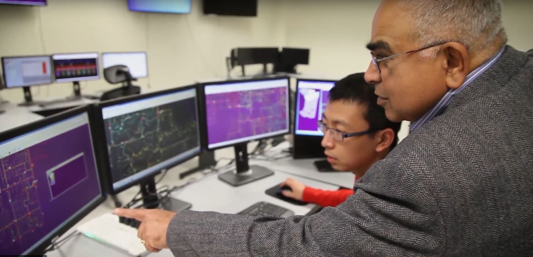 Researcher watches as Anjan Bose points at data on one of four computer monitors.