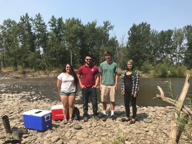 Four people standing next to ice chests by a river.