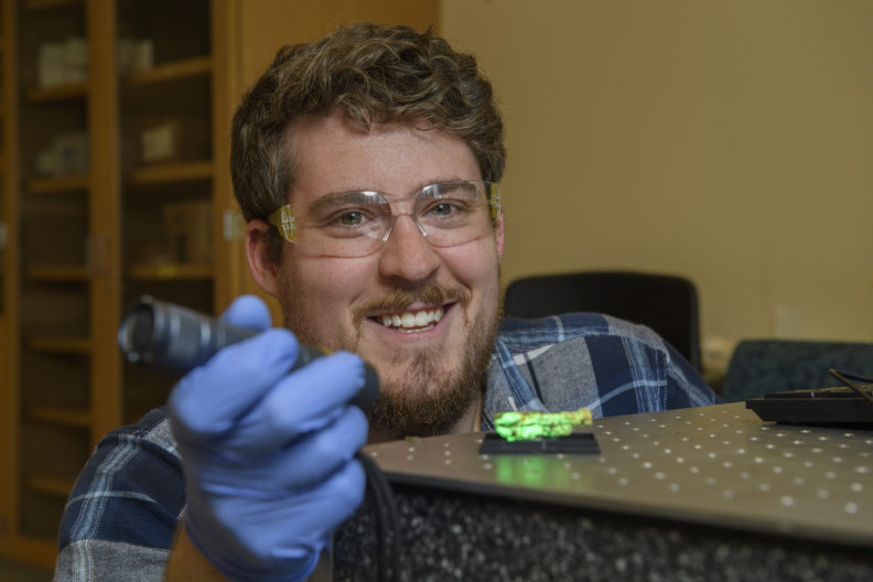 Travis Olds wearing safety glasses and gloves and shining a light on a mineral which glows green.