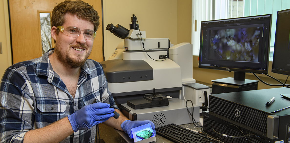 Travis Olds smiles at the camera while holding a rock sample at a computer station.