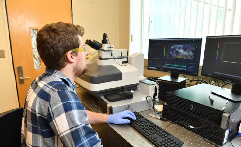 Travis Olds sitting at a computer workstation next to a microscope.
