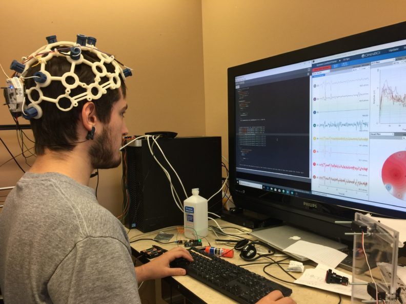 Alex Trevithick sitting in front of a computer monitor wearing a white helmet-like brain-computer interface. 