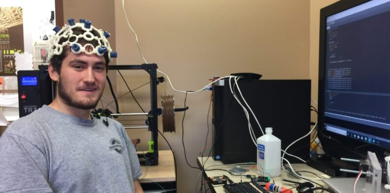 Alex Trevithick sits at a computer, wearing a helmet-like brain-computer interface, smiling at the camera.
