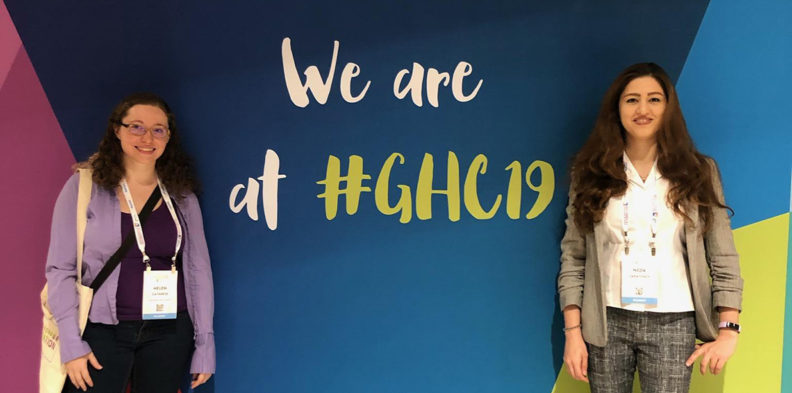 Two students pose in front of a backdrop with the text We are at #GHC19.