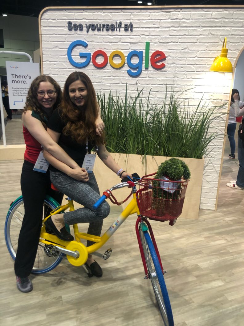 Two women pose on a bicycle by a display that says See Yourself at Google.
