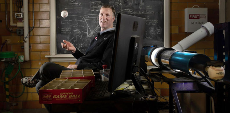 Lloyd Smith sits at a desk next to an air cannon while tossing a baseball in the air.
