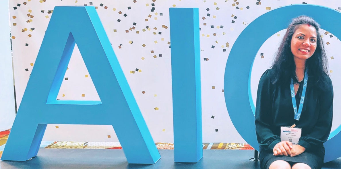 Neeru Chaudhary seated in front of "AIO" block letters.