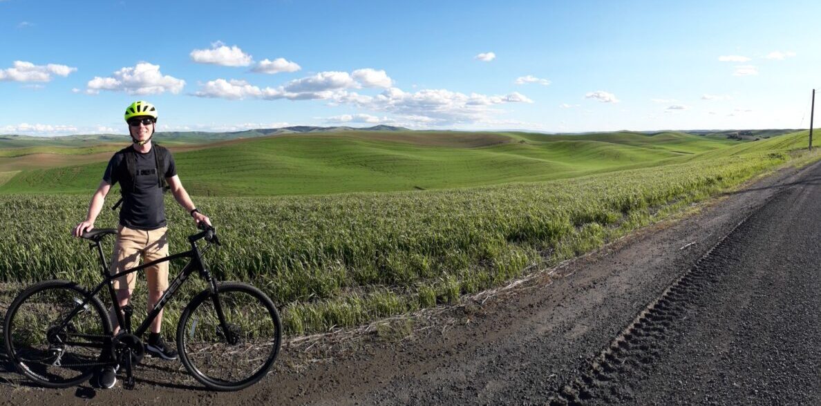 Andrew Raub standing by a bicycle against a backdrop of rolling green fields.