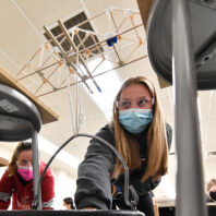 A model wooden truss bridge is balanced between two chairs with weight suspended beneath it.