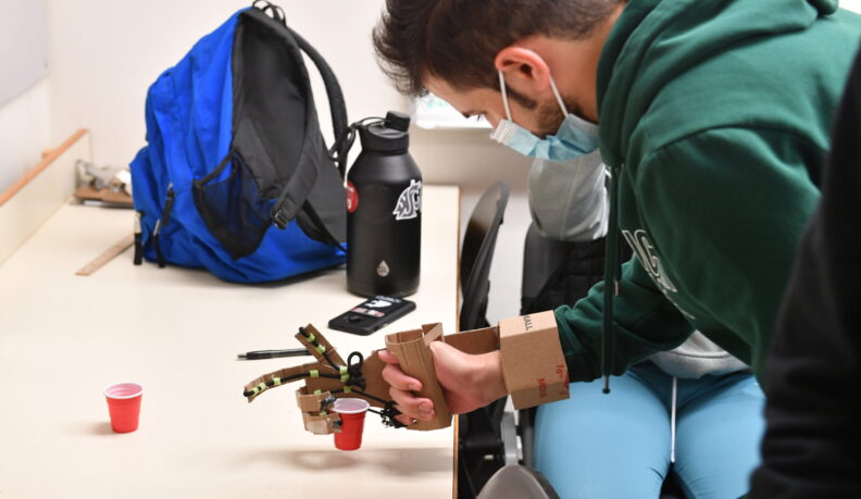 A student picks up a cup using a prosthetic hand made of cardboard and drawstring.