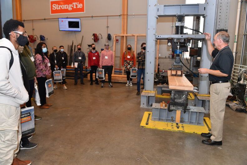 Don Bender stands next to machinery while addressing students.