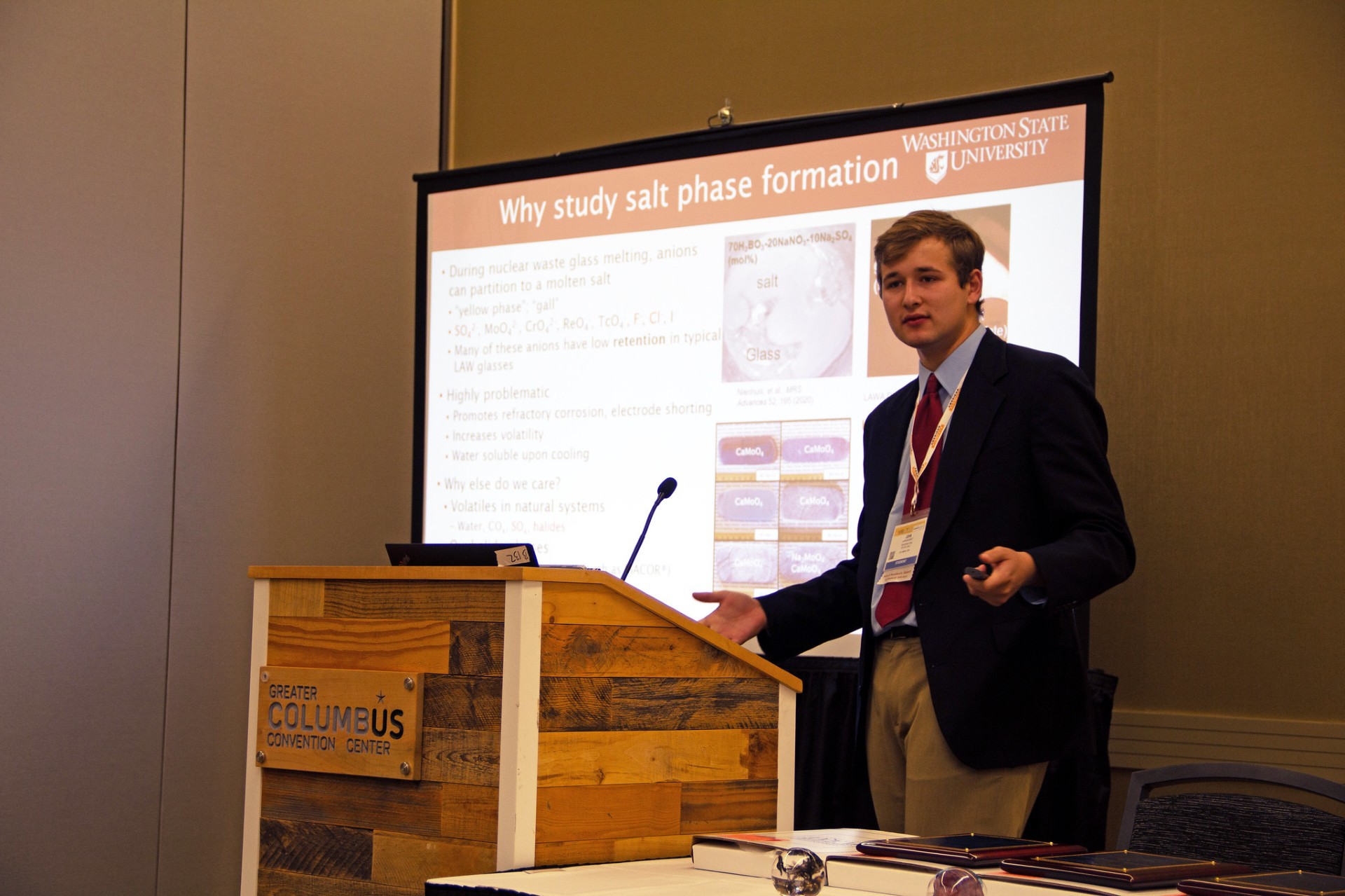 John Bussey speaking at a podium with a display screen that reads "why study salt phase formation" behind him.