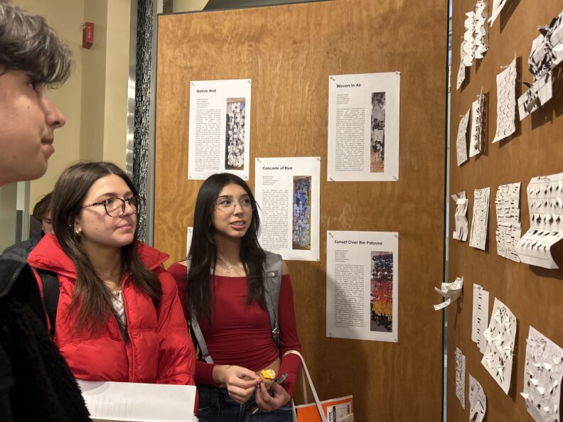 Three people looking at the white paper cut in intricate designs displayed on a hard board panel.