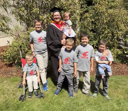 Brian Watson standing in their graduation cap and gown, standing beside their children.