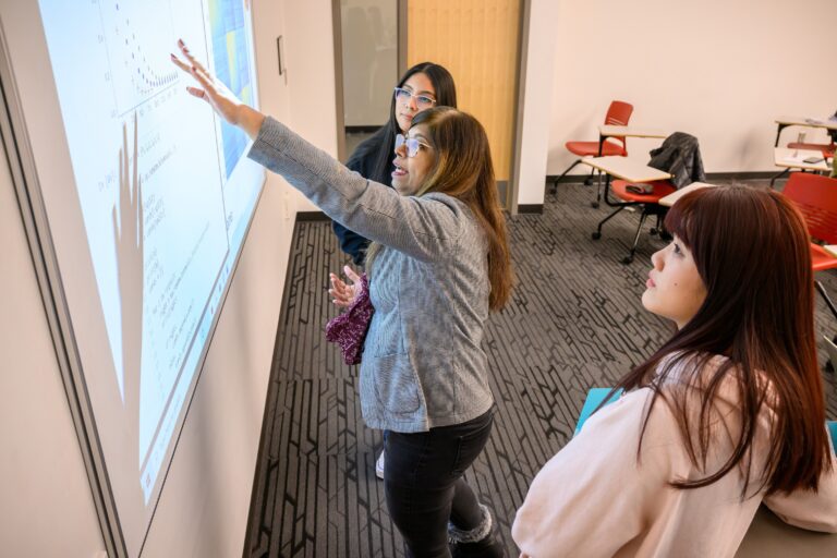 Professor gestures at projector screen in front of students in a classroom.
