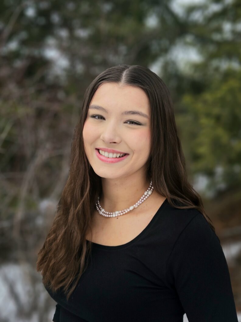 A smiling woman wearing a black top against a backdrop of trees.