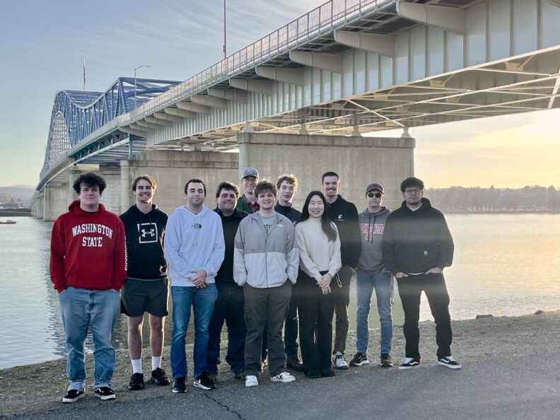 Students pose on the shore near a bridge spanning a river.