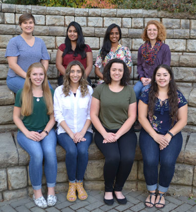 Eight women seated in two rows in front of a stone wall.