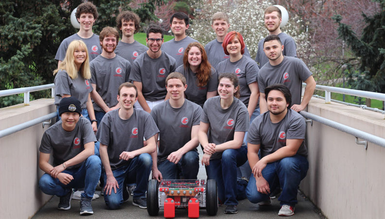 Sixteen robotics club members pose with a robot in front of Dana Hall in Pullman, WA.