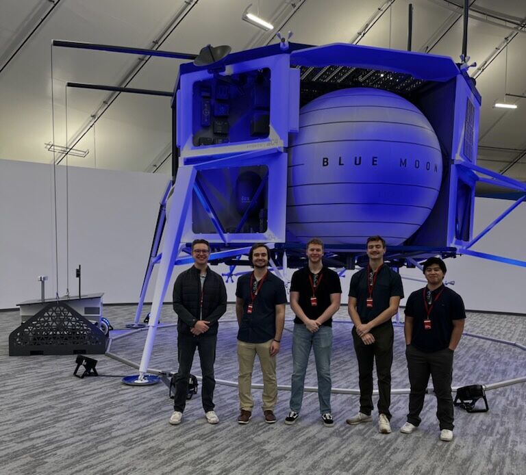Five students stand next to a model lunar lander.