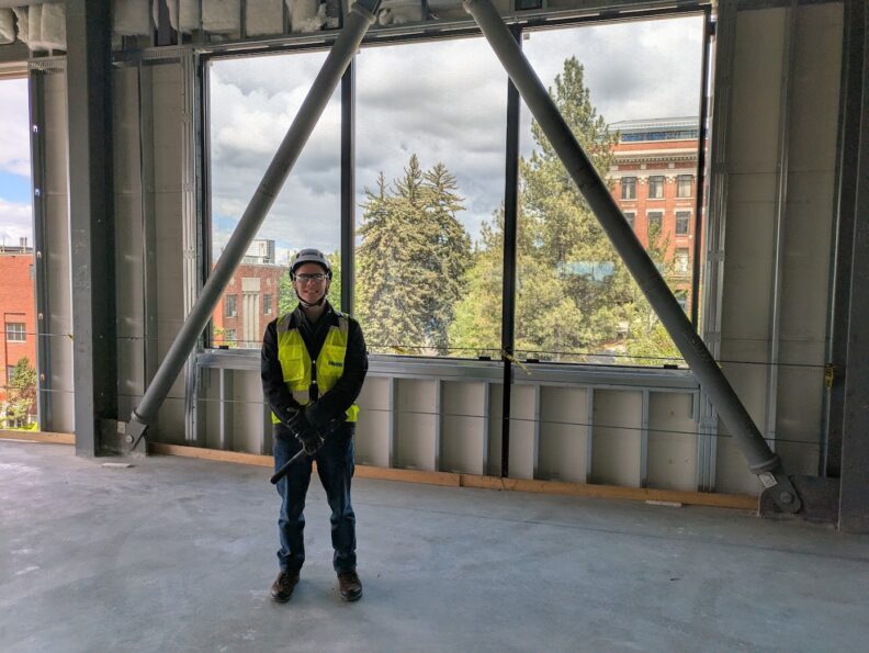 Camden wearing a hard hat, safety glasses, and reflective vest in a building under construction.