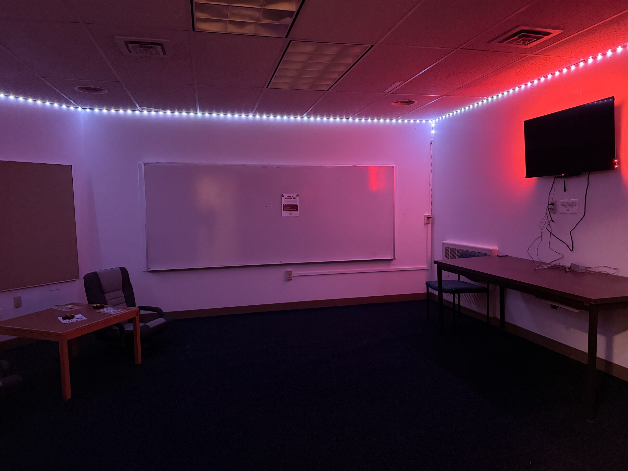 A large room with a television hanging on the right hand side and a large white board facing the camera. The room includes LED lights that changes colors across the perimeter of the room.