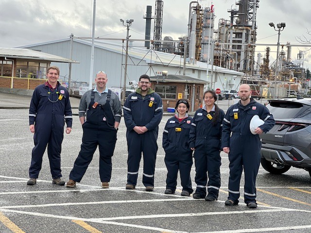 Six people in overalls with walkie-talkies pose in front of an industrial plant.