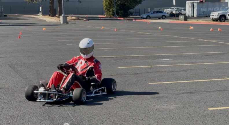 Man in a red racing suit driving a prototype in a parking lot.