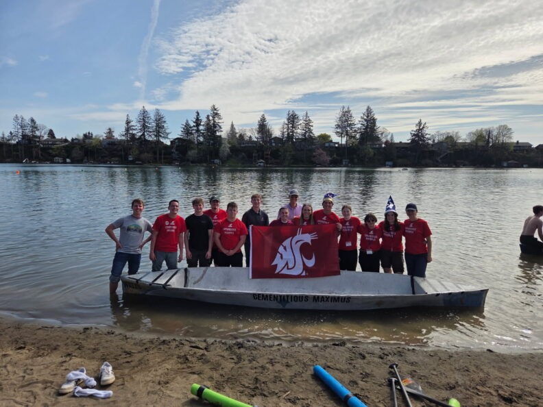 Students holding a WSU flag standing behind a concrete canoe parked in the water.