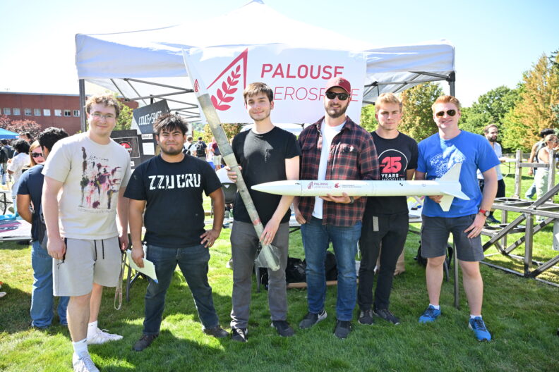 Six students stand at an outdoor booth displaying model rockets.