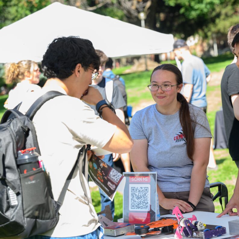 A student wearing a Crimson Robotics T-shirt speaks with another student at an outdoor campus event booth.