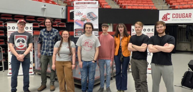 A group of sudents stand in line posing for a photo. Behind them is a poster for Crimson bot brawl.