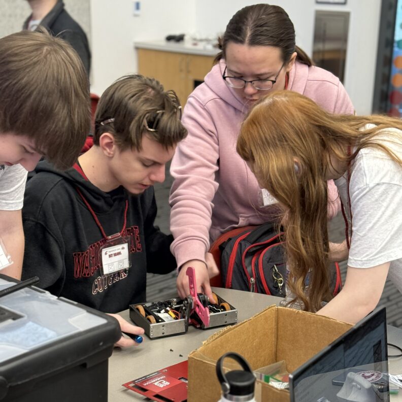 Four students gather around a small wheeled combat robot on a classroom table.