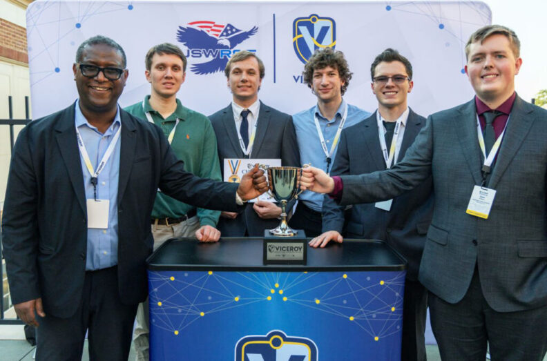 Professor and five students stand behind a table, smiling and holding a trophy. They wear event lanyards, and behind them is a backdrop with the logos ‘USWR’ featuring an eagle and ‘Viceroy.’ 