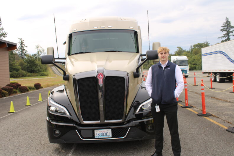 A person stands in front of a beige Kenworth semi-truck parked on a test track.