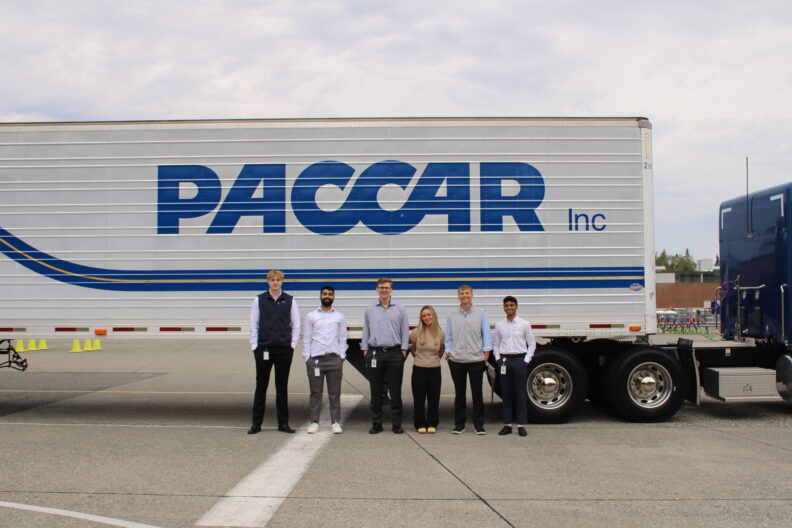 Six people stand in a row in front of a PACCAR semi-trailer displaying a large blue “PACCAR Inc” logo.