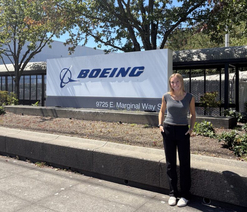 A person stands beside a large Boeing sign outside a facility, with trees and a clear blue sky in the background.