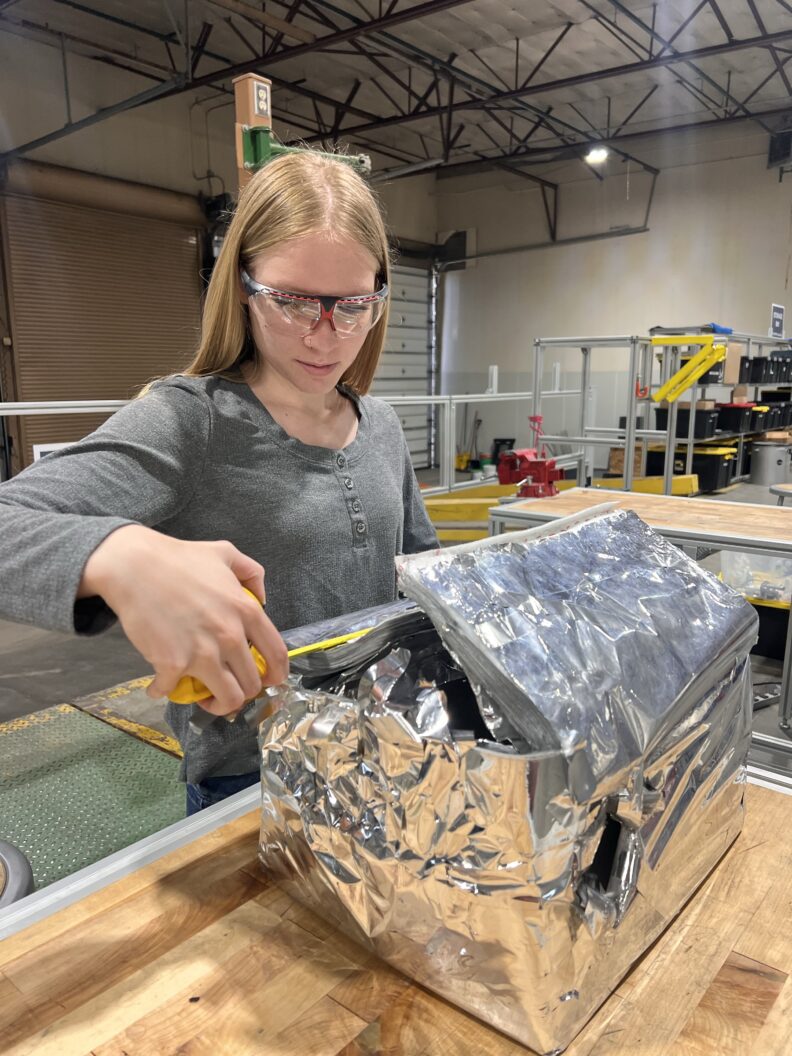 A person wearing safety glasses works on equipment covered with a multilayer insulation blanket in a workshop.