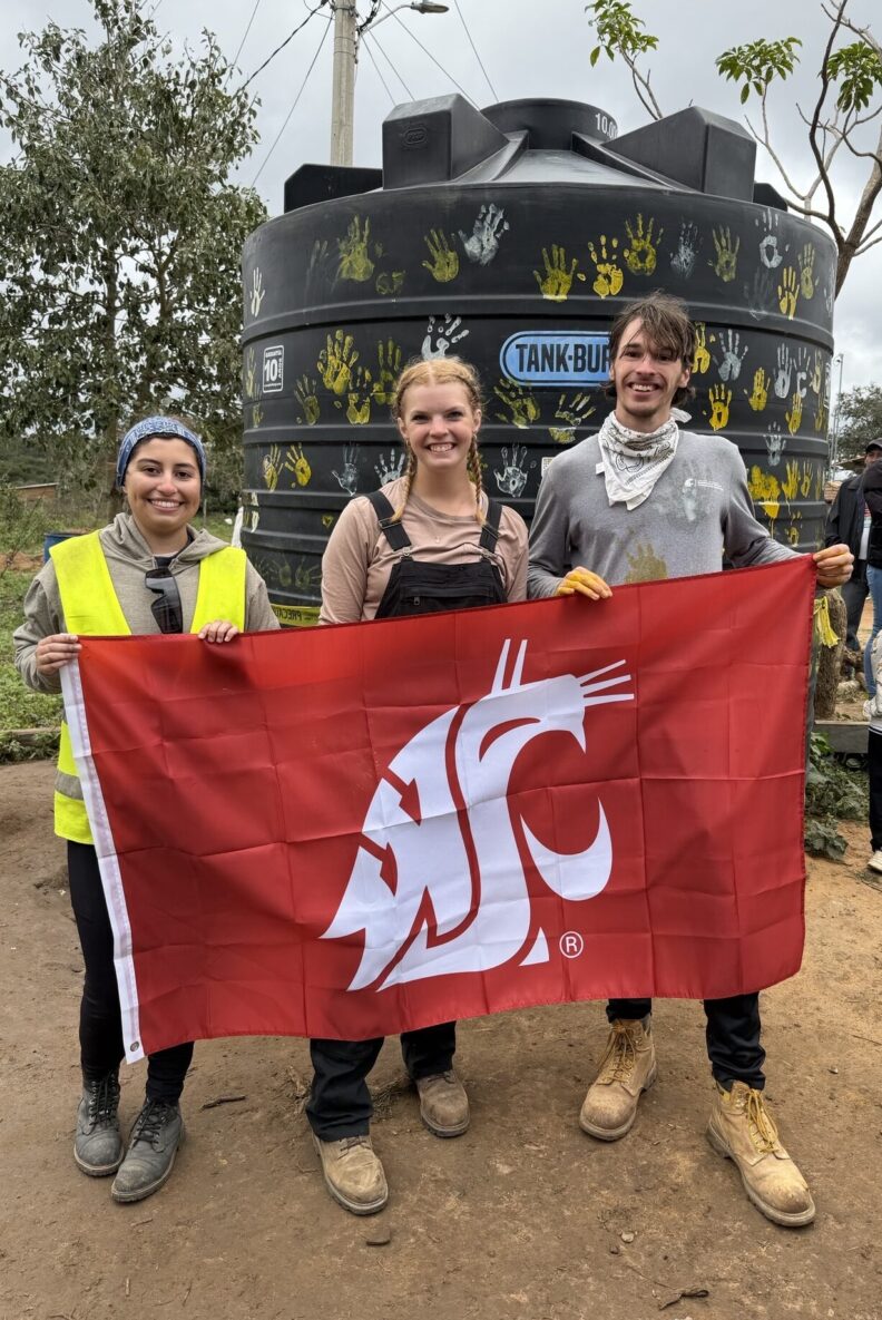 Three volunteers hold a red Washington State University flag in front of a large black water tank decorated with painted handprints.