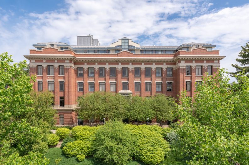 Four-story brick building with a fifth modern story added on top, surrounded by green landscaping.