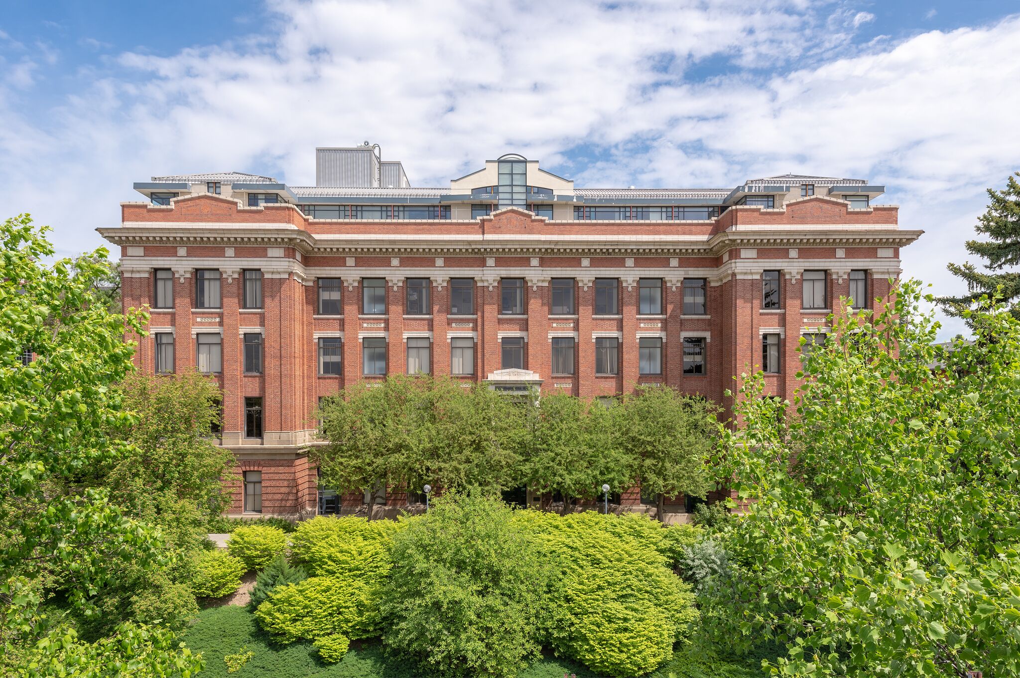 Four-story brick building with a fifth modern story added on top, surrounded by green landscaping.