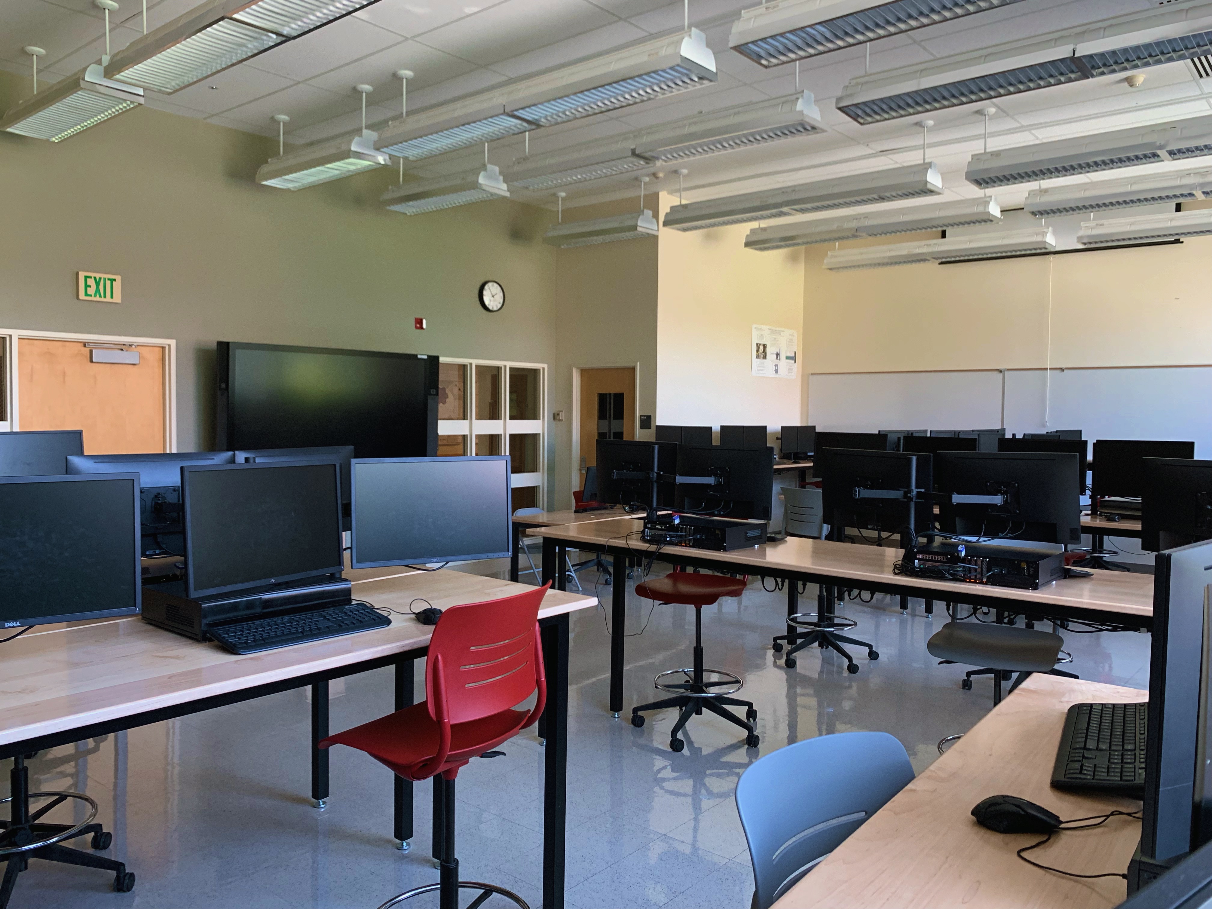 The room contains several dual-monitor computer stations on height-adjustable work tables and stool chairs as well as a SMART board in the front center of the room.