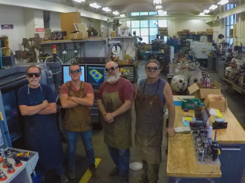 Four men standing in a row wearing safety goggles and aprons surrounded by shop equipment.