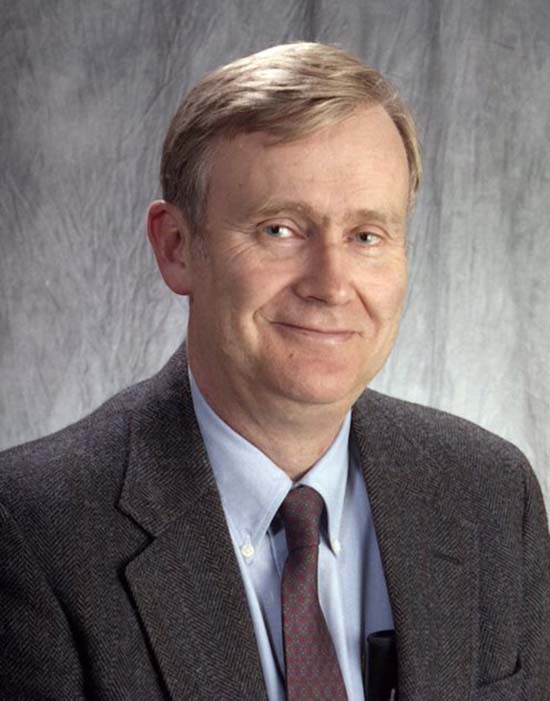 Dr. Wescott in a photo studio with a gray fabric background behind him. He is wearing a dark grey jacket with a shirt and tie.
