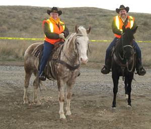 Two people on horseback. They are wearing orange safety vests.