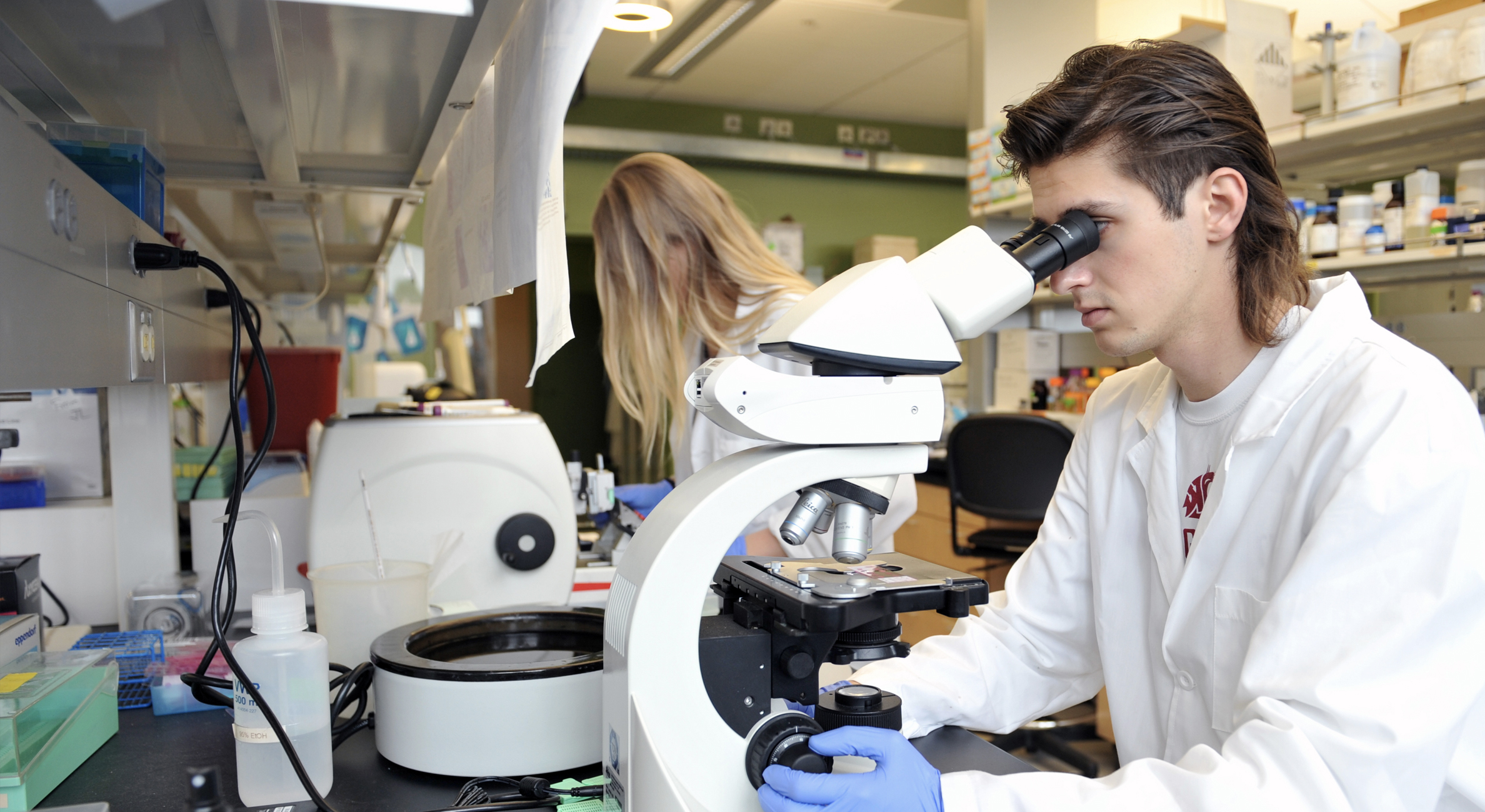 Two students in a lab. The one in the forefront is looking through a microscope.