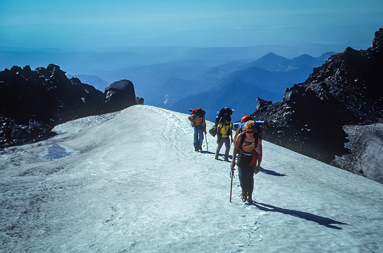 Hikers on a snow ridge with mountain ranges in the distance behind them.