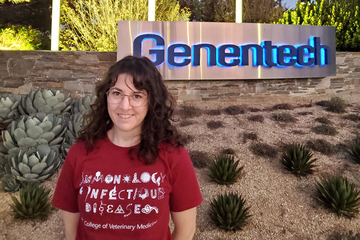 Alum, Jessica Klein, wearing a College of Veterinary Medicine shirt and standing in front of a Genentech sign.