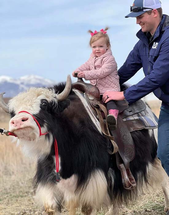 Henry holding his daughter as she's riding a saddled yak.