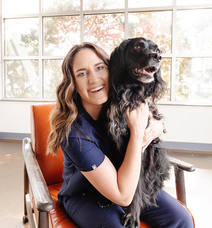 Jessica, sitting in an orange arm chair and holding a black spaniel in her lap.